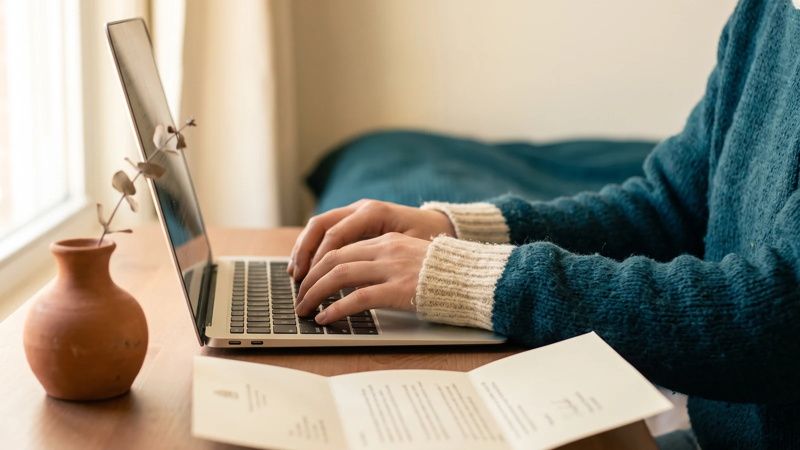 A person works at a laptop at a desk by a window, papers and a small vase beside them